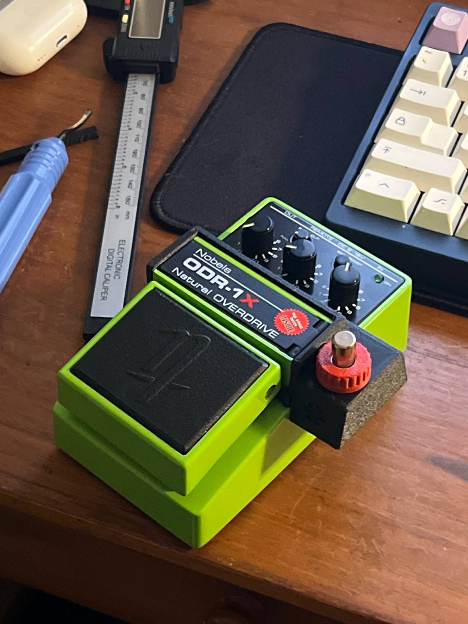 A Nobels ODR-1X with a green Cltchworks NB-1 footswitch fitted, on a workbench beside digital calipers and a screwdriver